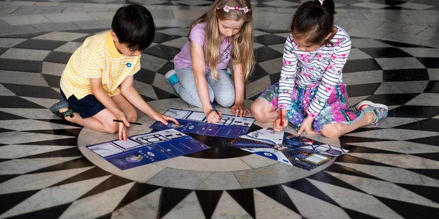 Children fill out an activity sheet during a visit to the Queen's House in Greenwich. They are sat on the floor, at the centre of an intricate black and white marble pattern